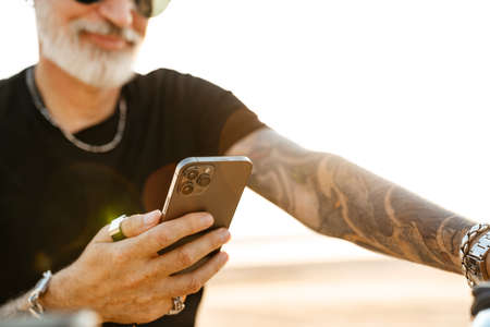 Mature bold man with tattoo using cellphone while sitting on motorbike outdoorsの写真素材
