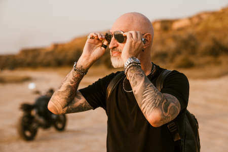Bold senior man wearing eyeglasses posing with motorcycle outdoors on summer day