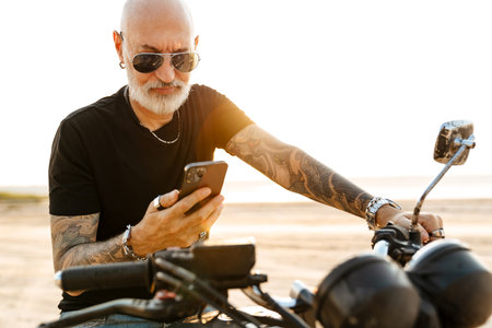 Mature bold man with tattoo using cellphone while sitting on motorbike outdoorsの写真素材