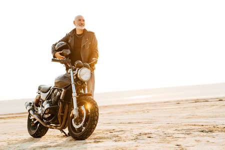 Bold senior man in leather jacket posing with motorcycle outdoors on summer day