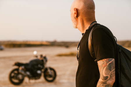 Bold senior man wearing t-shirt posing with motorcycle outdoors on summer dayの写真素材