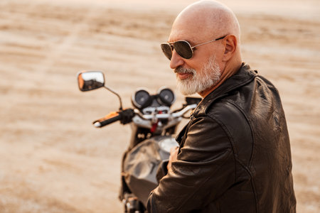 Bold senior man wearing leather jacket posing on motorcycle outdoors on summer day