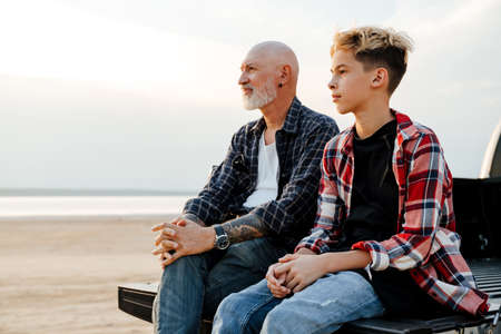 Smiling mid aged father and his preschooler son sitting on a back of a car parked at the beach...