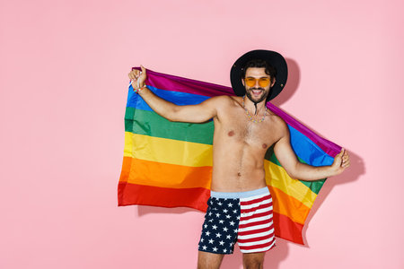 Shirtless man in hat laughing while posing with rainbow flag isolated over pink wallの写真素材