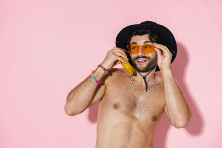 Shirtless man in hat looking aside while posing with banana isolated over pink wall
