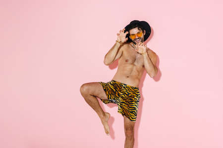 Shirtless man in hat laughing and gesturing at camera isolated over pink wall