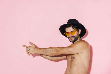 Shirtless man in hat laughing and making gun gesture isolated over pink wall