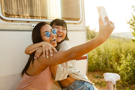 Multiracial two women hugging while taking selfie on cellphone by trailer outdoorsの写真素材