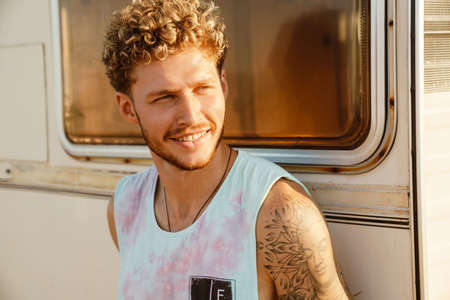 Ginger young man smiling and looking aside while leaning on trailer outdoors