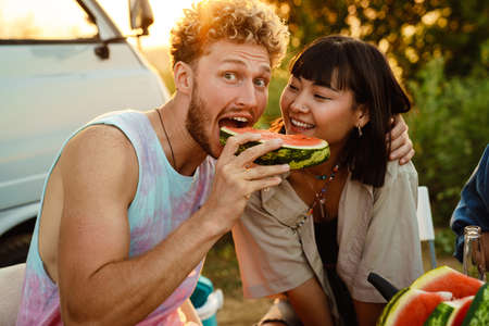 Multiracial couple eating watermelon during picnic with their friends by trailer outdoorsの写真素材