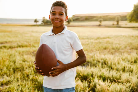 Black boy wearing white shirt smiling while posing with rugby ball outdoors