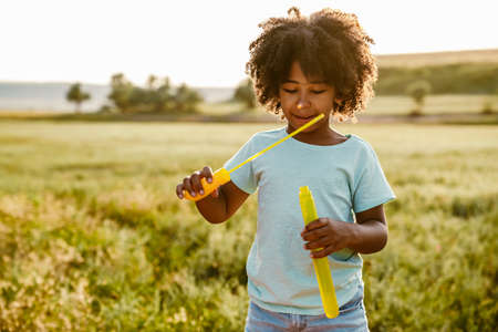 Black curly boy wearing t-shirt smiling while making soap bubble outdoors