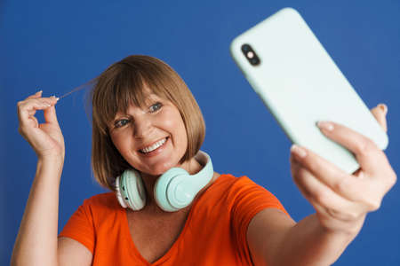 Senior blonde woman with headphones taking selfie on mobile phone isolated over blue wallの写真素材