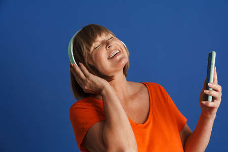 Senior blonde woman smiling while listening music with headphones isolated over blue wall