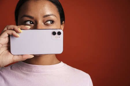 Young black woman wearing t-shirt posing with cellphone isolated over red wall