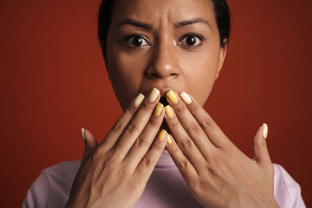 Young black woman covering her mouth while expressing surprise at camera isolated over red wall