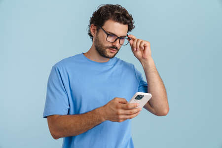Young brunette man in eyeglasses using mobile phone isolated over blue background