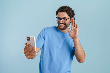 Young brunette man gesturing while using mobile phone isolated over blue backgroundの写真素材