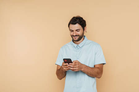 White bristle man wearing shirt smiling and using mobile phone isolated over beige background