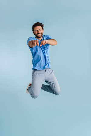 Young brunette man jumping while pointing fingers at camera isolated over blue background