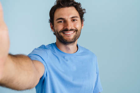 Young brunette man with bristle smiling while taking selfie photo isolated over blue background