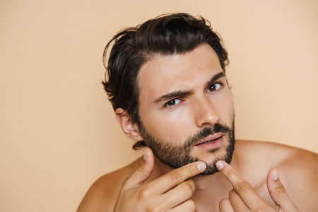 Young half-naked man squeezing pimple on his face isolated over beige backgroundの写真素材