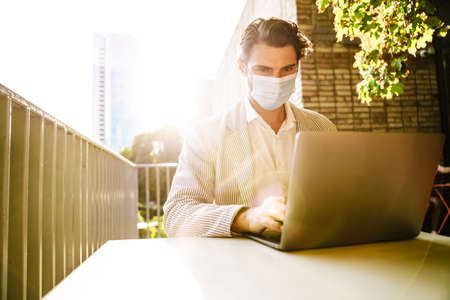 Young man in face mask working with laptop while sitting at cafe outdoors