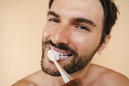 Young half-naked man smiling while brushing his teeth isolated over beige backgroundの写真素材