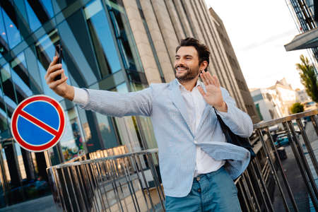 Young man gesturing while taking selfie photo on mobile phone outdoors