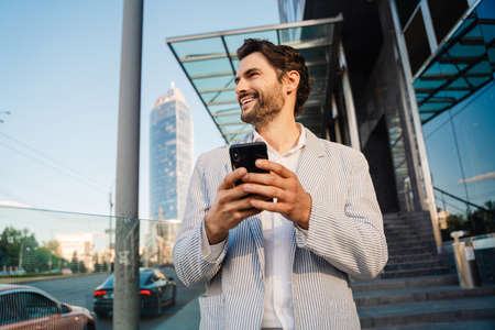 Young man wearing jacket smiling and using mobile phone outdoorsの写真素材