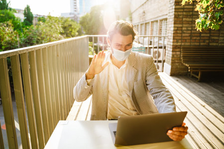 Young man in face mask gesturing while working with laptop at cafe outdoorsの写真素材