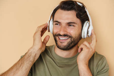 Young white man smiling while listening music with headphones isolated over beige background