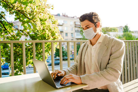 Young man in face mask working with laptop while sitting at cafe outdoorsの写真素材