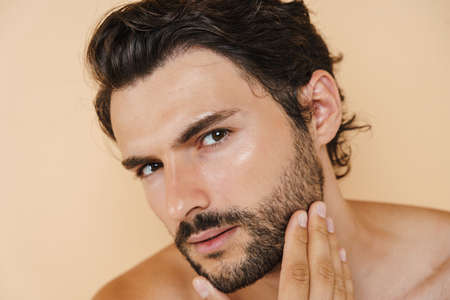 Young half-naked man rubbing his bristle and looking at camera isolated over beige background