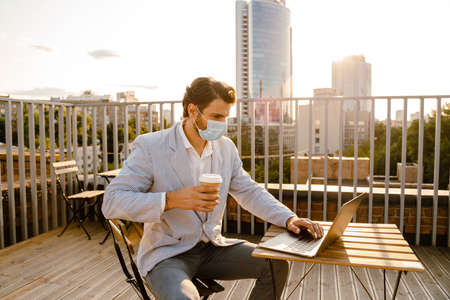 Young man in face mask working with laptop while sitting at cafe outdoorsの写真素材