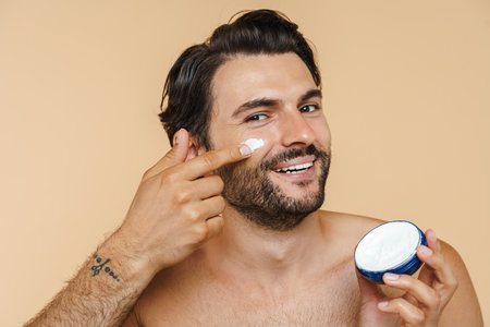 Young half-naked man smiling while applying face cream isolated over beige backgroundの写真素材
