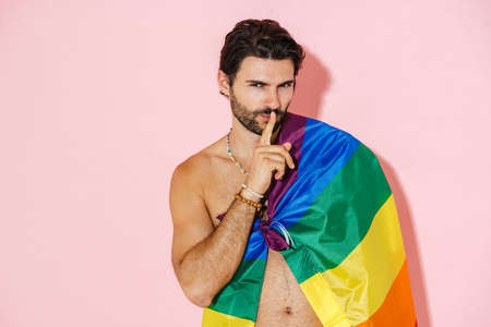 Young shirtless man with rainbow flag showing silence gesture isolated over pink backgroundの写真素材