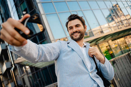 Young man smiling while taking selfie photo on mobile phone outdoorsの写真素材
