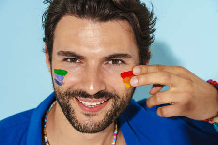 Young man wearing blue jacket smiling while doing rainbow makeup isolated over blue background