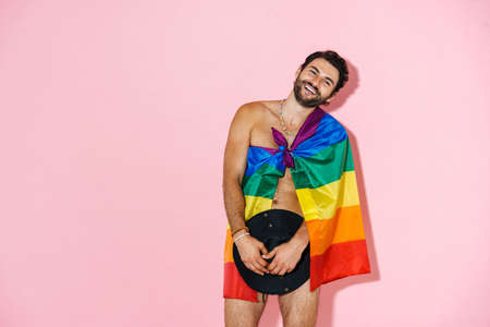 Young naked man laughing while posing with rainbow flag and hat isolated over pink backgroundの写真素材