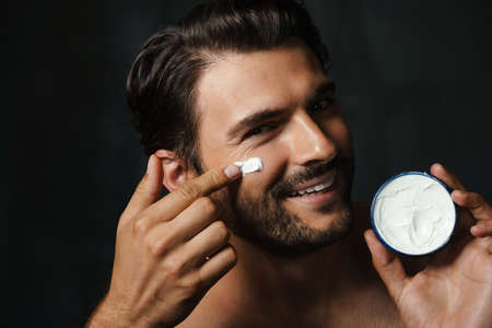 Young half-naked man smiling while applying face cream isolated over black backgroundの写真素材