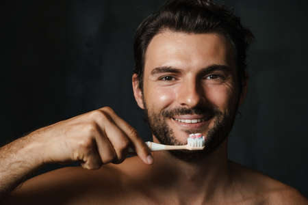 Young half-naked man smiling while brushing his teeth isolated over black backgroundの写真素材