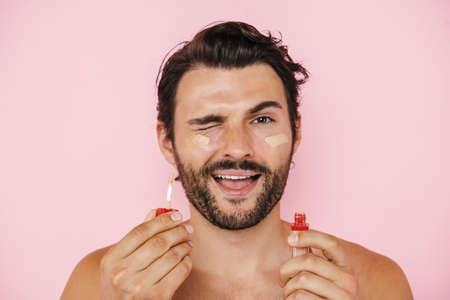 White shirtless man winking while applying concealer isolated over pink backgroundの写真素材