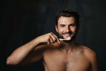 Young half-naked man smiling while brushing his teeth isolated over black backgroundの写真素材