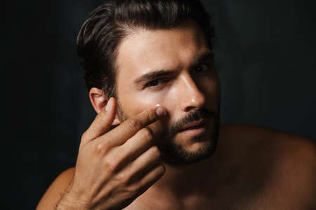 Young half-naked man looking at camera while applying face cream isolated over black backgroundの写真素材