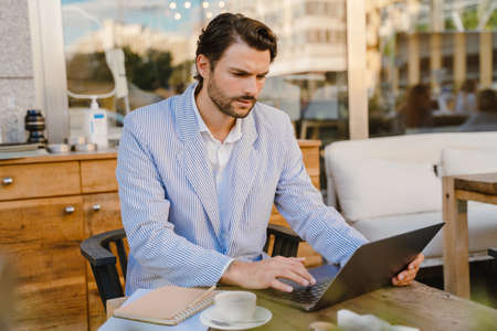 Young man wearing jacket working with laptop while sitting at cafe outdoors