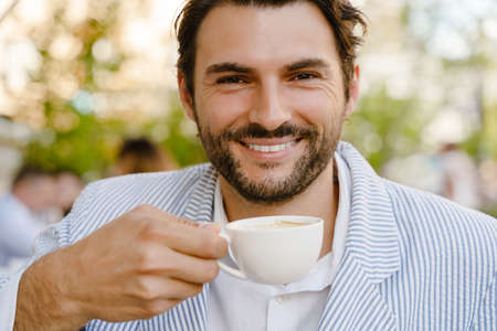 Young brunette man smiling and drinking coffee while sitting at cafe outdoorsの写真素材