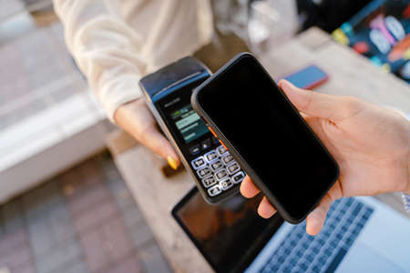 Young man paying cellphone in cafe while waitress holding payment terminalの写真素材