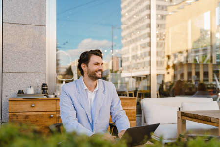 Young man wearing jacket working with laptop while sitting at cafe outdoorsの写真素材