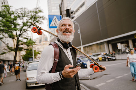 Grey mature man using mobile phone while standing with skateboard at city street